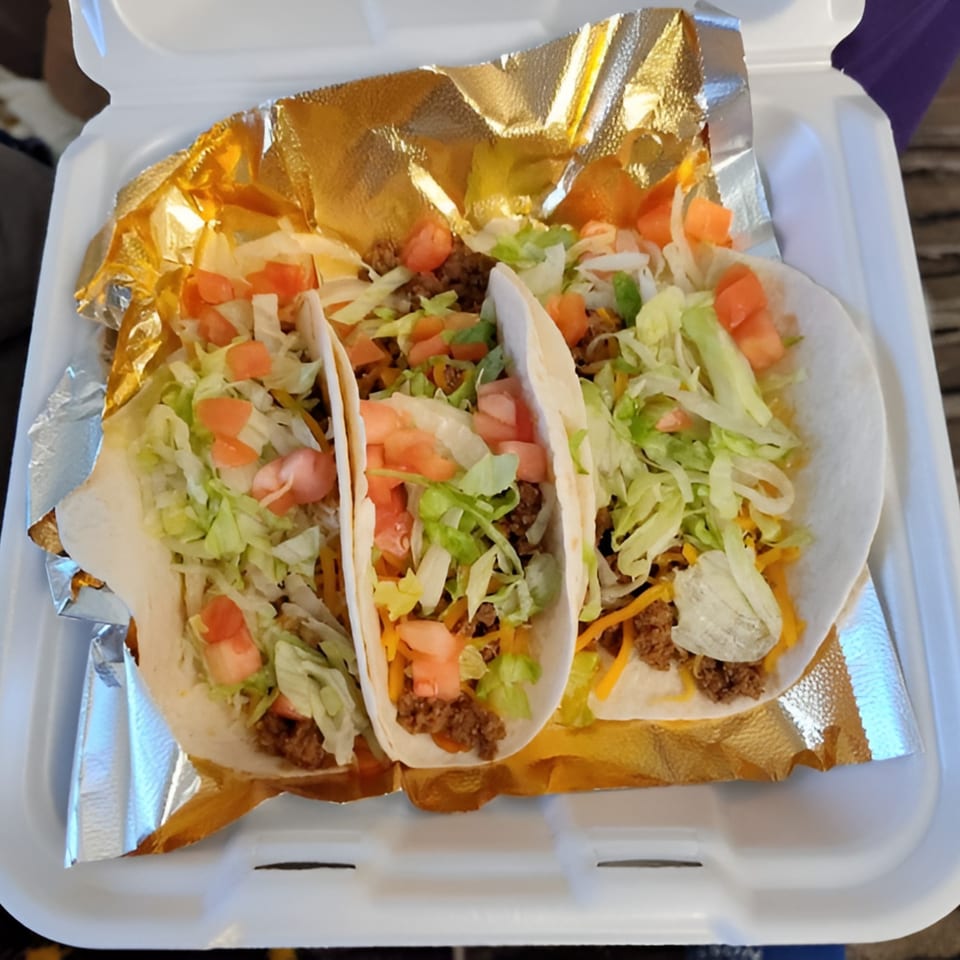 Three soft tacos with ground beef, shredded lettuce, diced tomatoes, and cheese in a takeout container.