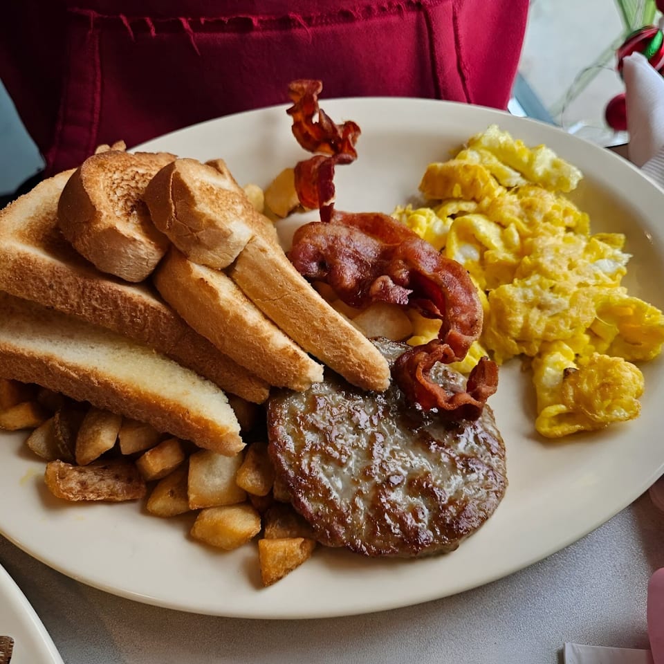A diner breakfast plate with scrambled eggs, bacon, sausage patty, home fries, and toast.