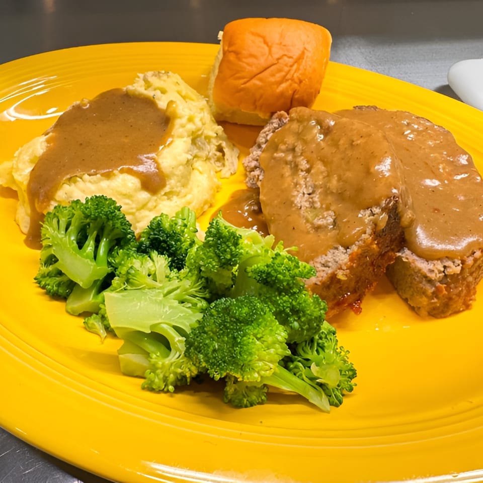 Meatloaf with gravy, mashed potatoes, broccoli, and a dinner roll on a bright yellow plate.